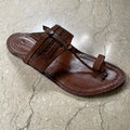 A single brown leather kolhapuri sandal with a decorative woven strap and a small metal charm, shown from an overhead angle on a textured concrete floor.