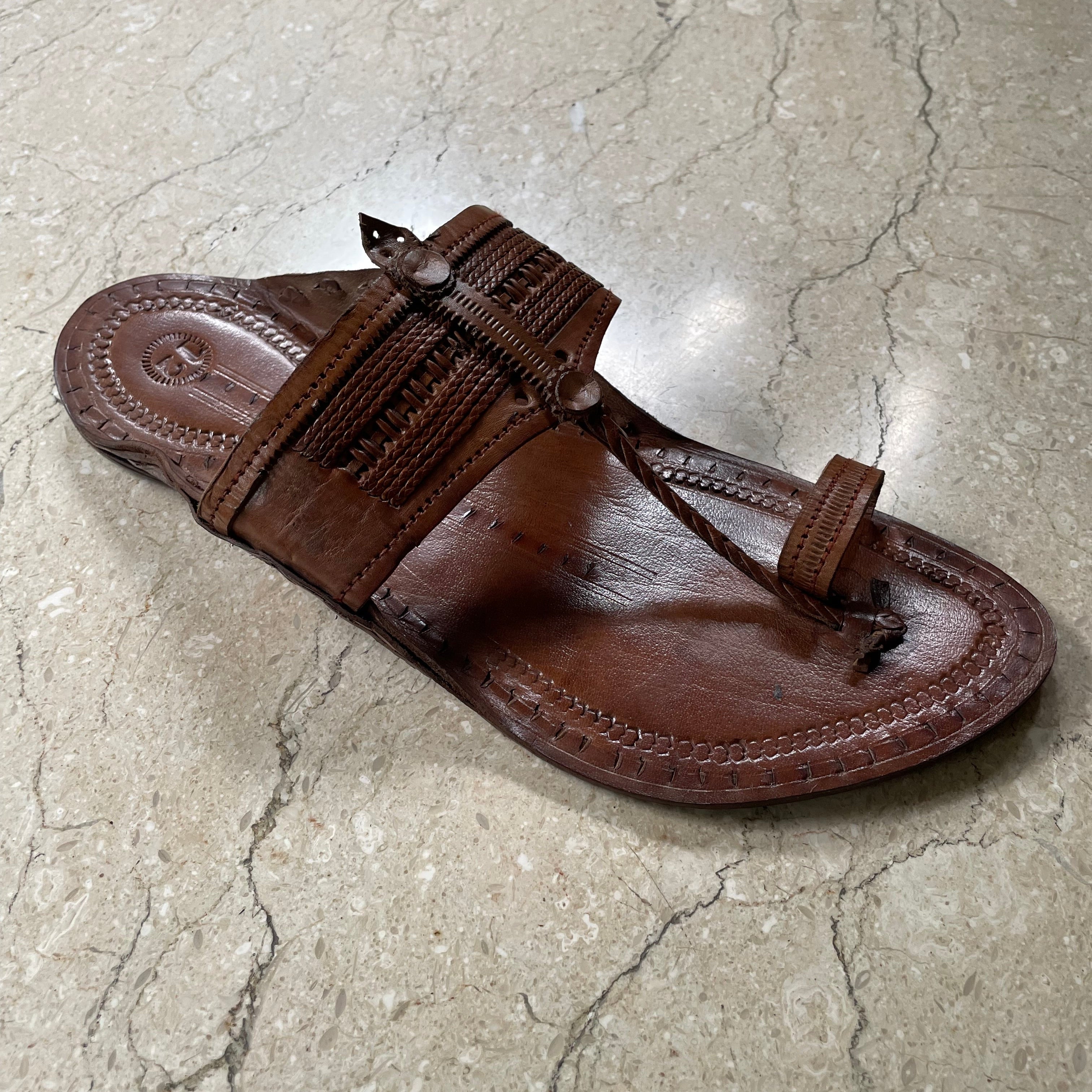 A single brown leather kolhapuri sandal with a decorative woven strap and a small metal charm, shown from an overhead angle on a textured concrete floor.