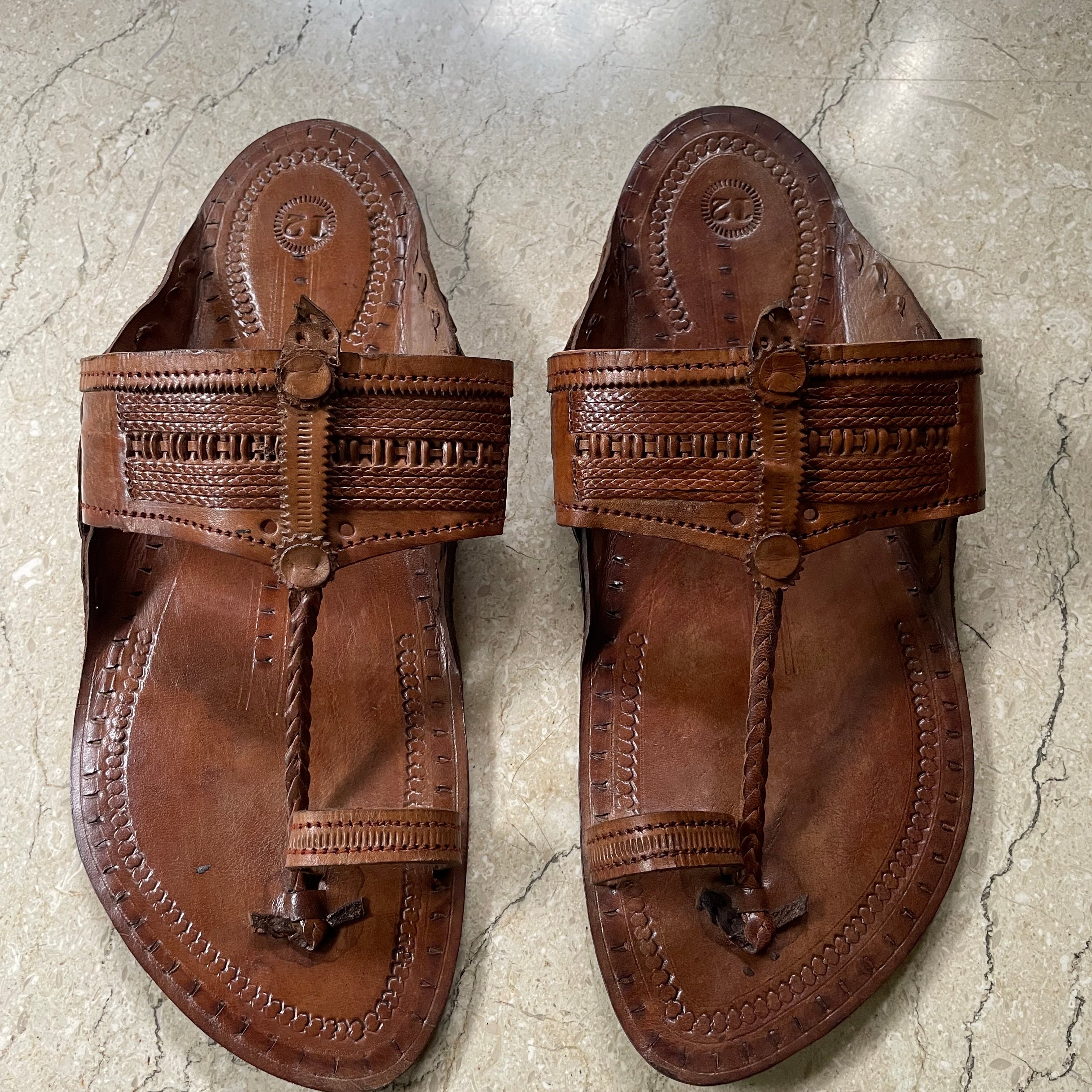A pair of brown leather kolhapuri  sandals with intricate braided and stitched details, displayed flat on a light gray stone floor.