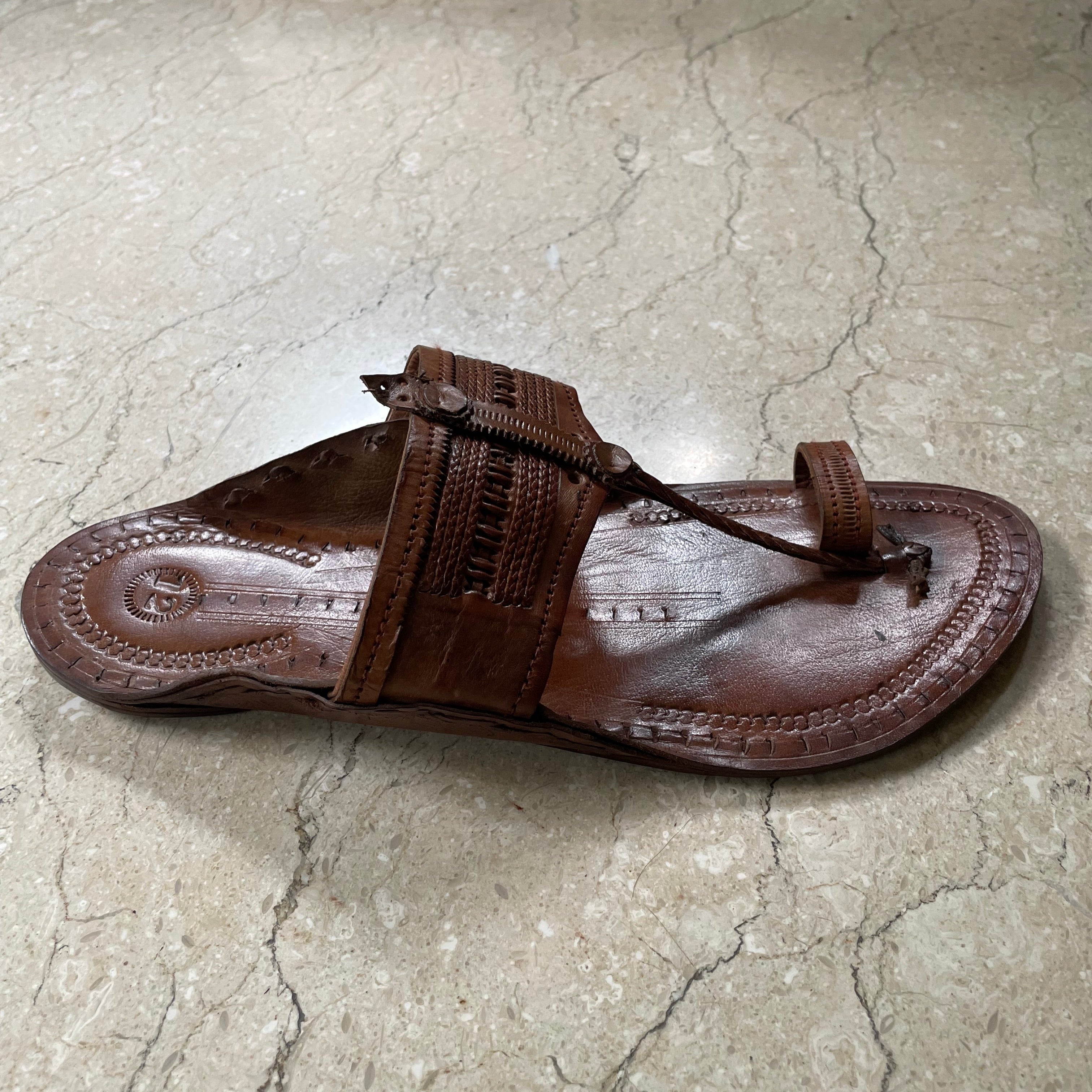 A single brown leather kolhapuri sandal with a woven toe strap, photographed on a light gray stone-tiled floor next to a cardboard box.
