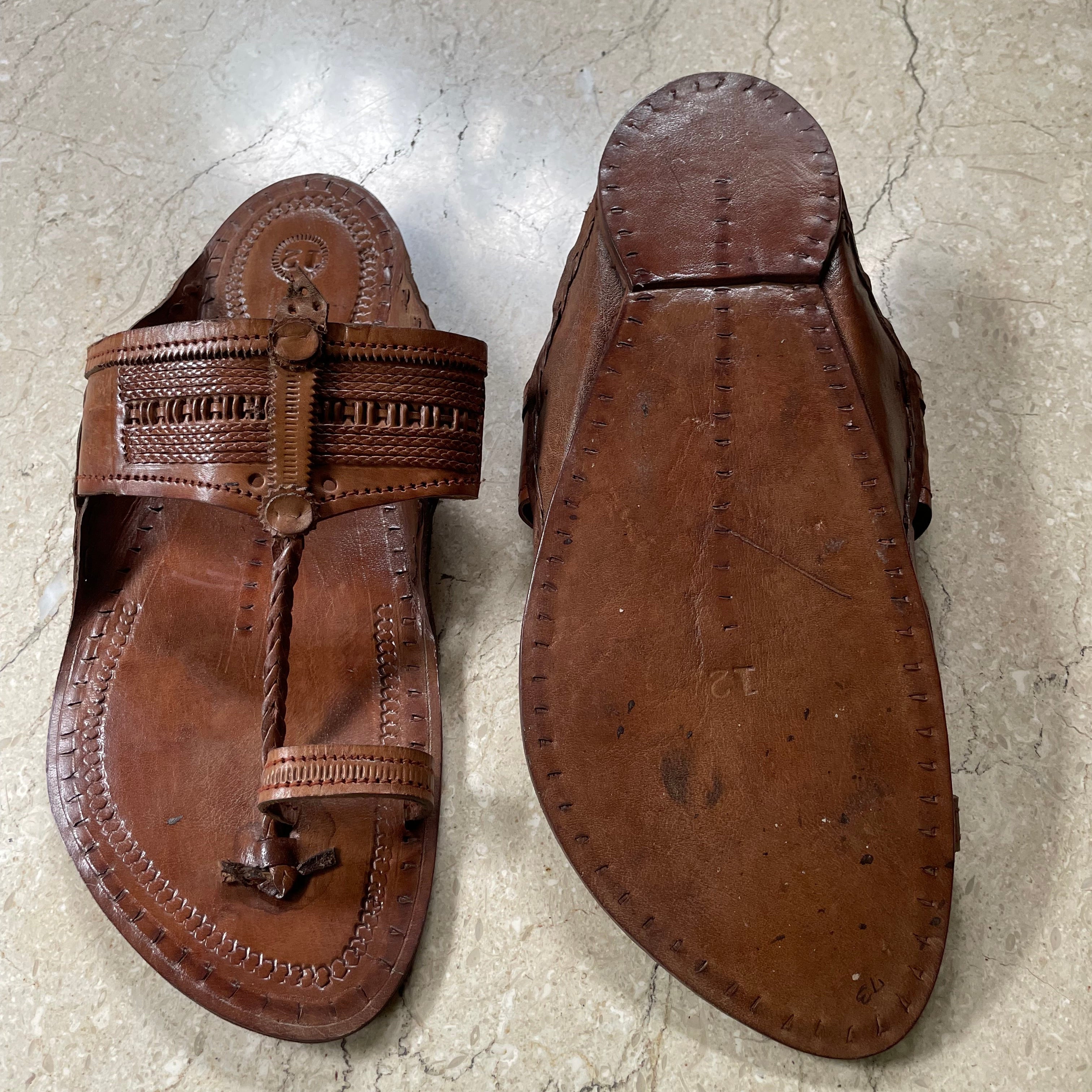 A pair of brown leather kolhapuri  sandals shown from above on a light stone floor, displaying the sole and the front strap with decorative stitching and a metal ring.