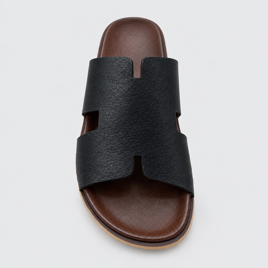 A single black slide sandal with a wide cutout strap and a brown footbed, photographed from above on a beige marble floor.
