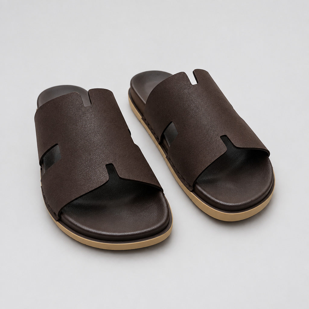 A pair of dark brown leather slide sandals with a cutout design, resting on a polished beige marble floor.